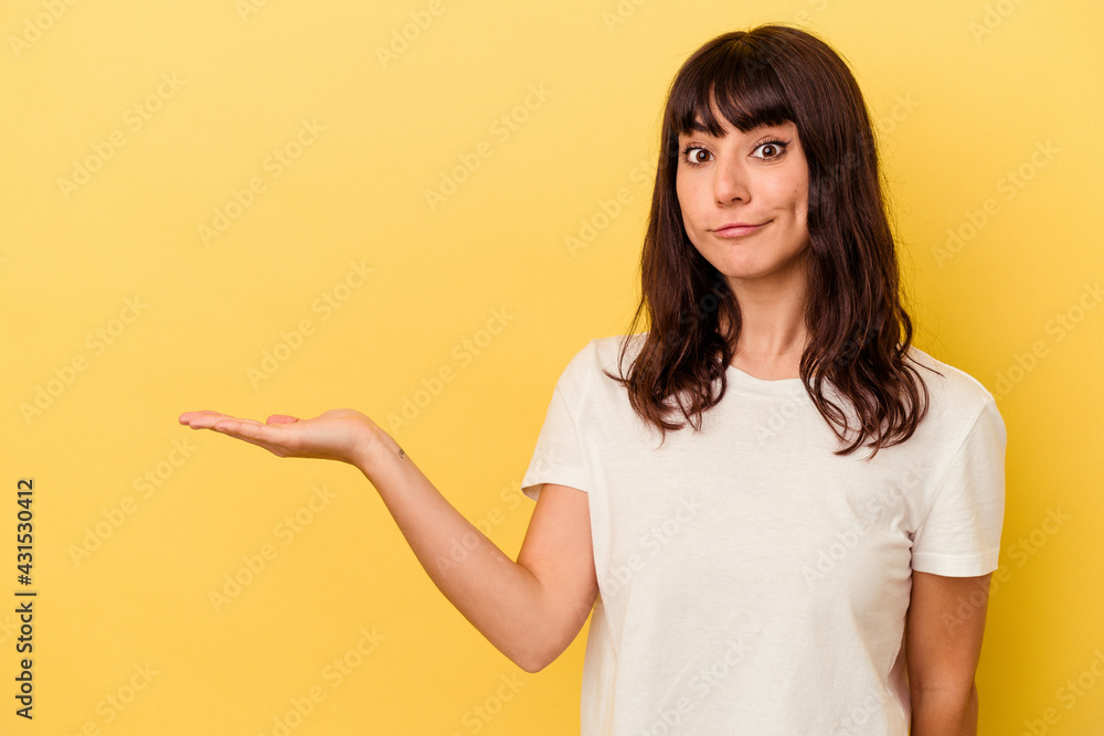 Young caucasian woman isolated on yellow background showing a copy space on a palm and holding another hand on waist. Young caucasian woman isolated on yellow background showing a copy space on a palm and holding another hand on waist.