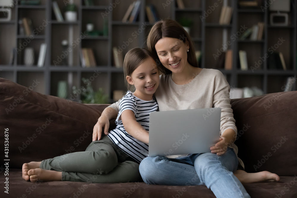Loving smiling mother and small daughter sit relax on sofa at home use laptop together talk on video call. Happy Caucasian mom and little teen daughter have fun study learn online on computer gadget. Loving smiling mother and small daughter sit relax on sofa at home use laptop together talk on video call. Happy Caucasian mom and little teen daughter have fun study learn online on computer gadget.