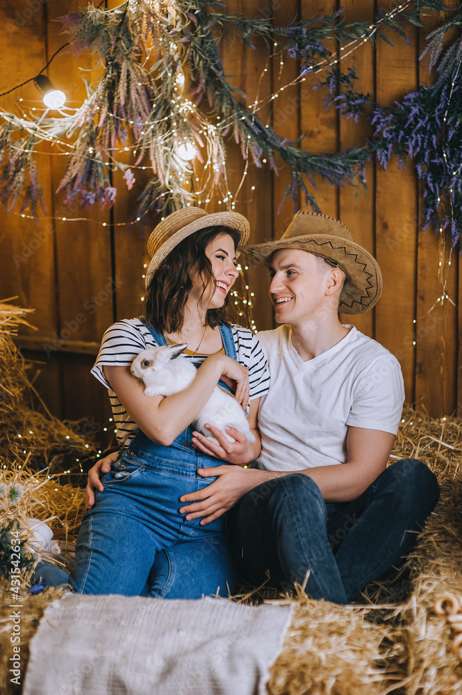 A loving man cowboy and a pregnant woman in a straw hat are sitting in the hay with a white rabbit in their arms, smiling and hugging in the countryside. Photography, concept.