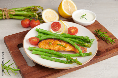 Delicious seasonal green asparagus and sliced smoked salmon on a rustic plate with lemon, salt, pepper, of lemon on a white plate . Baked salmon garnished with asparagus and tomatoes with herbs.