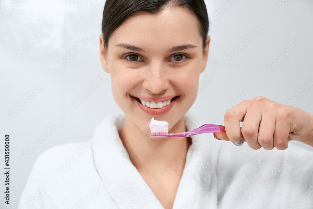 Close up portrait of smiling young beautiful woman in white robe after shower holding toothbrush with toothpaste. Concept of process preparing for cleaning teeth in morning. Close up portrait of smiling young beautiful woman in white robe after shower holding toothbrush with toothpaste. Concept of process preparing for cleaning teeth in morning.