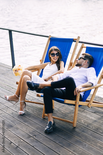 Beautiful newlyweds are relaxing on sun loungers, talking, smiling, against the background of the water. © Ivan