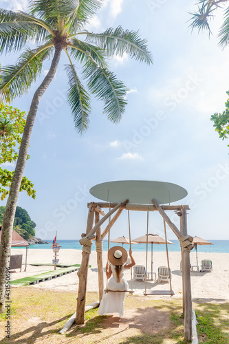 Back View of Travel Woman in White Dress and Straw Hat Sitting on Wooden Swing, Tropical Palms and Sandy Beach with Blue Sea on Background. Female Tourist Leisure in Resort on Phuket Island, Thailand © TravelMedia