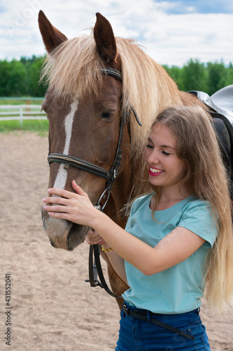 Portrait of smiling young woman with blond long hair is caressing brown horse snoot with big nostrils. Horseback riding hobby, vacation in countryside.