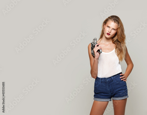 Front image of a young woman blonde posing in white fashion clothes in studio, looking at camera, holding a retro camera in hand. Copy space.