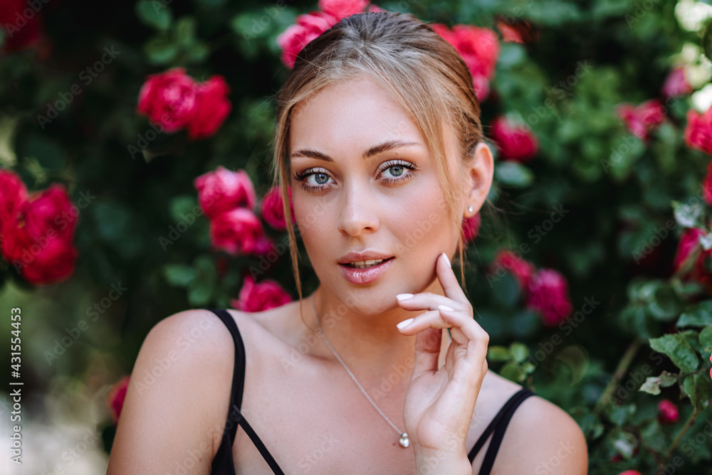 Outdoor photo of romantic young woman in rose garden. girl with red lips in a black dress . Young model on a background of a bush of roses. Stylish woman.