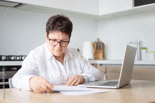 Adult senior 60s woman calculating paper documents at home at pc. Serious middle aged woman at home working on bank payments online with computer technologies.