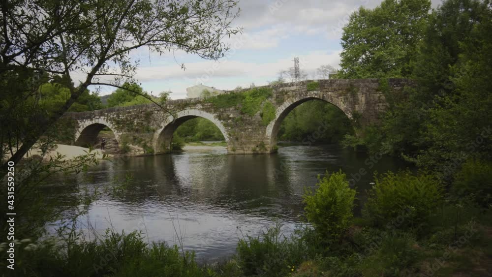 Roman stone bridge that crosses a river. Roman bridge of Ponteareas. stone bridge with green vegetation, cloudy sky and water with reflections, with beach on the shore Roman stone bridge that crosses a river. Roman bridge of Ponteareas. stone bridge with green vegetation, cloudy sky and water with reflections, with beach on the shore