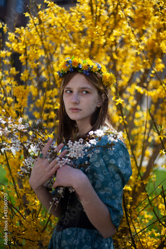 Girl in a blue dress in a wreath with yellow dandelions on his head on a background of yellow flowering bush