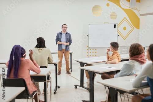 Wide angle view at mature male teacher giving lecture to class of young students in modern school, copy space