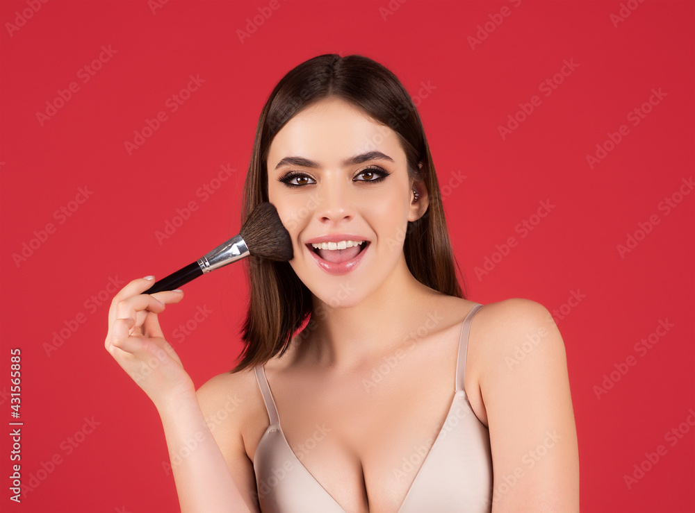 Beauty portrait of beautiful female model on red. Facial makeup. Young female model putting blush with cosmetic brush. Woman with beauty face, natural skin, facial skin portrait. Beauty portrait of beautiful female model on red. Facial makeup. Young female model putting blush with cosmetic brush. Woman with beauty face, natural skin, facial skin portrait.