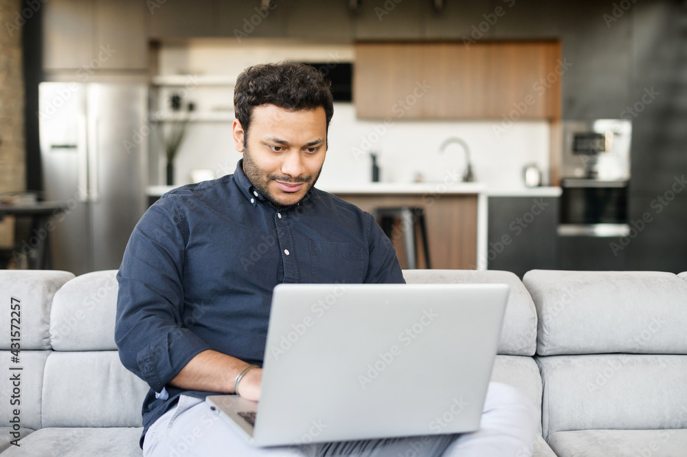 Smiling cheerful mixed-race freelancer guy is using laptop computer sitting on the couch at home, hindu guy enjoys remote work from home in comfy atmosphere. Indian male spends leisure online Smiling cheerful mixed-race freelancer guy is using laptop computer sitting on the couch at home, hindu guy enjoys remote work from home in comfy atmosphere. Indian male spends leisure online