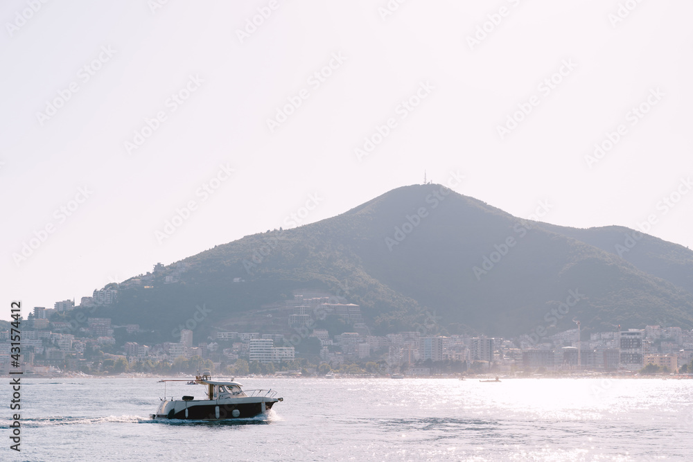 A pleasure boat with a sun awning on the coastline against the backdrop of the mountains and the city of Budva in Montenegro.