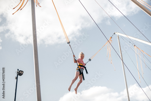 A girl of about 8 years old is jumping on a bungee trampoline. A child with a harness and elastic bands hangs against the sky. The concept of a happy childhood and games in an amusement park by the se