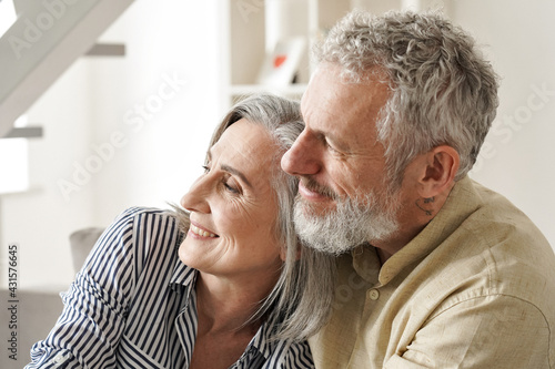 Happy older mature classy couple hugging, bonding, thinking of good future. Carefree mid age couple looking away dreaming together, enjoying comfort and wellbeing, feeling hope and pleasure at home. photo