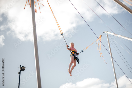 A girl of about 8 years old is jumping on a bungee trampoline. A child with a harness and elastic bands hangs against the sky. The concept of a happy childhood and games in an amusement park by the se