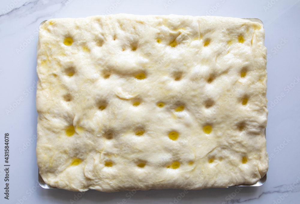 Traditional focaccia dough made from flour, water, yeast, sugar, salt water, and olive oil holes - top view, cooking process on a baking sheet on a marble table