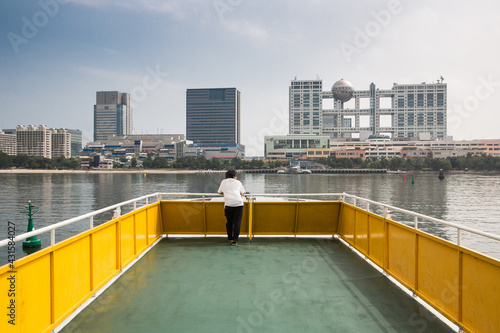 Lone Japanese man alone on boat. Odaiba water ferry iwith Odaiba Island and Aqua City in the distance. Tokyo, Japan photo