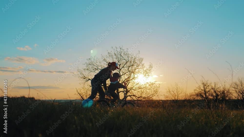 Childhood bike concept. Mother teaching son to ride bicycle. Happy cute boy learn to riding a bike in park at sunset time. Young mom teaching son to ride bike first time on countryside rural road. Childhood bike concept. Mother teaching son to ride bicycle. Happy cute boy learn to riding a bike in park at sunset time. Young mom teaching son to ride bike first time on countryside rural road.