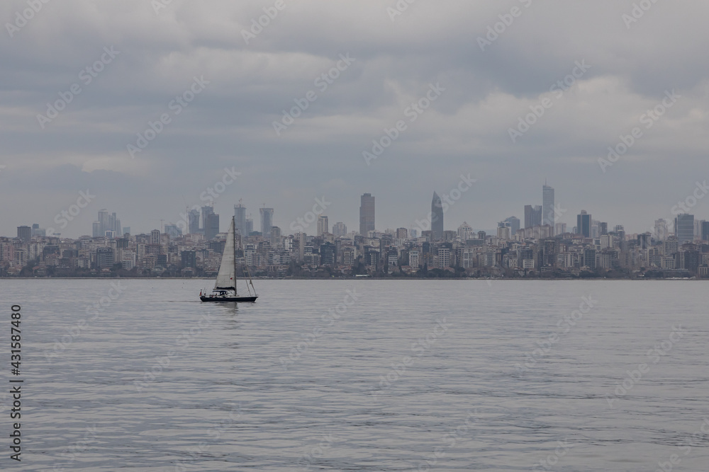 A small lonely boat is floating under the sail in the wide sea space against the backdrop of the gloomy faded city line of Istanbul along the coast. Cloudy dull day at the sea.