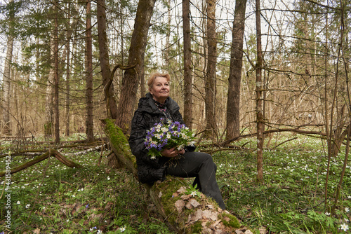 An elderly woman of 65 years with a bouquet of spring flowers sits on a fallen tree in the forest.