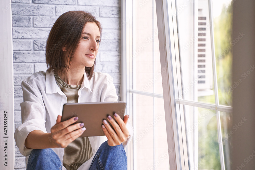 Young woman sitting at windowsill. Working on tablet. Online study concept. Safety lockdown distance teaching. Female person. Quarantine office. Business writer. Video relax. Copyspace Young woman sitting at windowsill. Working on tablet. Online study concept. Safety lockdown distance teaching. Female person. Quarantine office. Business writer. Video relax. Copyspace