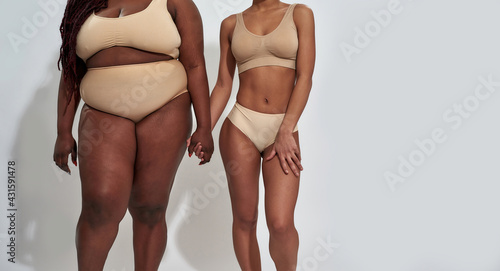 Cropped shot of two african american women in underwear with different body size holding each other hands, standing together isolated over gray background