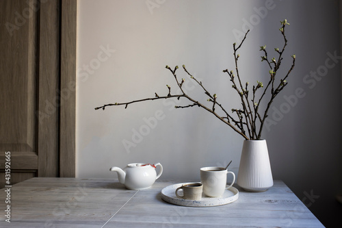 Half-open branches of an apple tree in a white fluted vase. A kettle and two cups of coffee on a tray on the table.