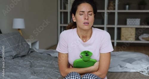 Woman in pajamas sit on bed in bedroom suffer from dysmenorrhea painful period menstrual cramps hug hand warmer hot water bag to reduce ache. Unhealthy female, digestion problem, need medicare concept photo