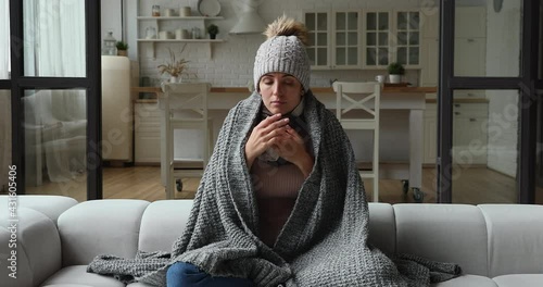 Young woman wear hat wrapped in knitted plaid sit alone shivering from cold on sofa in unheated apartment without central heating due debt. Unhealthy female feeling discomfort try to warming up indoor