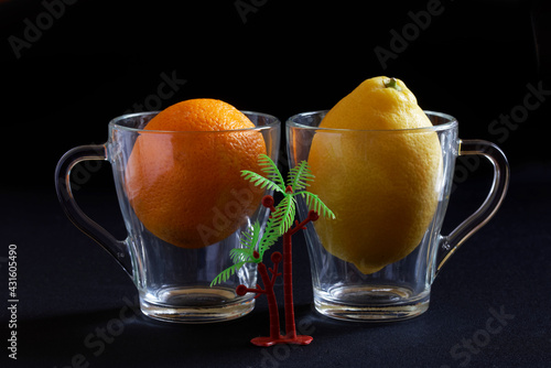Fresh fruits - lemon and orange in transparent glass mugs next to a toy palm tree. Tropical juice concept