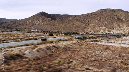 Drone following a black SUV driving over a road in the middle of a dry grassland, beautiful mountain scenery in the background, Murica, Spain. photo