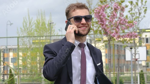 Young Caucasian man in suit, outside, spring time, smiling, talking on phone with sunglasses