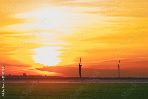 many windmills stand on a large field spinning in the evening against a sunset orange sky. Alternative Energy Concept