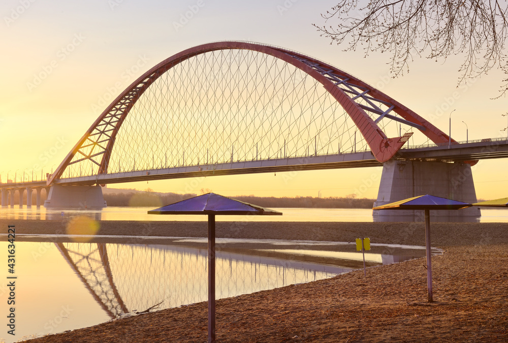 Naklejka premium Dawn of the Bugrinsky Bridge. A deserted beach in spring, an arched modern road bridge over the Ob River in golden light. Novosibirsk, Siberia, Russia