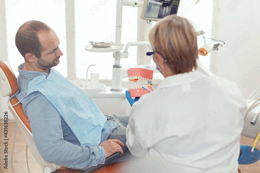 Stomatologist holding sanitary model of the mandible talking with sick man. Senior dentist explaining correct dental hygiene using mock-up skeleton of teeth while working in stomatology hospital Stomatologist holding sanitary model of the mandible talking with sick man. Senior dentist explaining correct dental hygiene using mock-up skeleton of teeth while working in stomatology hospital