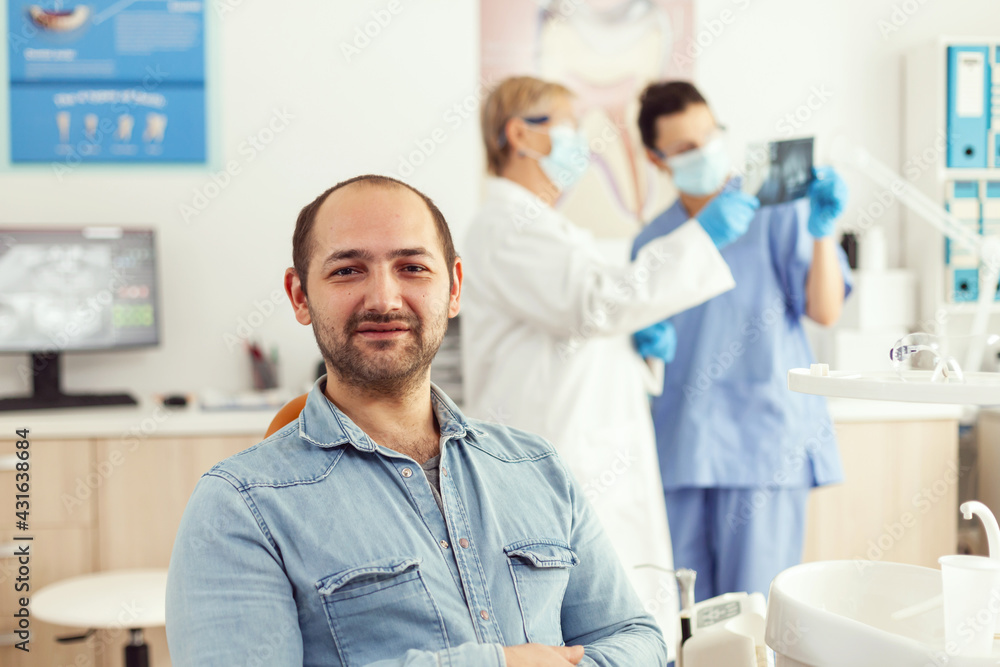 Fototapeta premium Man patient looking into camera waiting for radiography results sitting on dental chair in hospital stomatology clinic. In background dentists team looking at tooth x-ray preparing for surgery