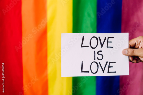 love is love written on white paper held by a hand with a lgbt flag in the background.