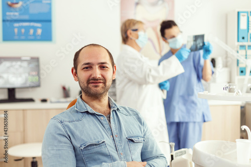 Man patient looking into camera waiting for radiography results sitting on dental chair in hospital stomatology clinic. In background dentists team looking at tooth x-ray preparing for surgery
