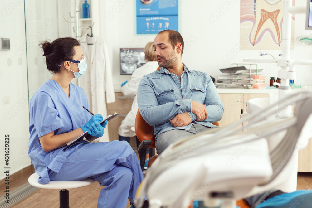 Obraz premium Orthodontist nurse discussing with man patient about tooth consultation writing healthcare treatment on clipboard sitting in medical dental office. Stomatologist making surgery appointment