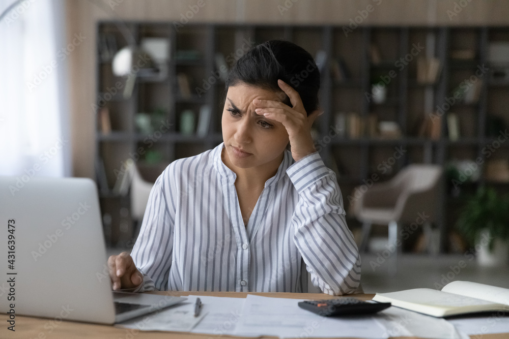 Fototapeta premium Frustrated Indian woman calculating bills, looking at laptop screen, touching forehead, unhappy young female planning managing budget, using internet banking service, checking financial documents