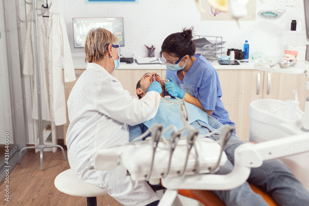 Senior woman dentist analyzing teeth examining sick patient while sitting on orthodontic chair. Doctor stomatologist and nurse cleaning mouth during dental examination in stomatology clinic Senior woman dentist analyzing teeth examining sick patient while sitting on orthodontic chair. Doctor stomatologist and nurse cleaning mouth during dental examination in stomatology clinic