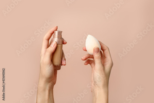 Applying foundation on makeup sponge. Woman's hands with neutral manicure holding bottle of concealer or toner foundation, cream and beauty blender, make up artist background