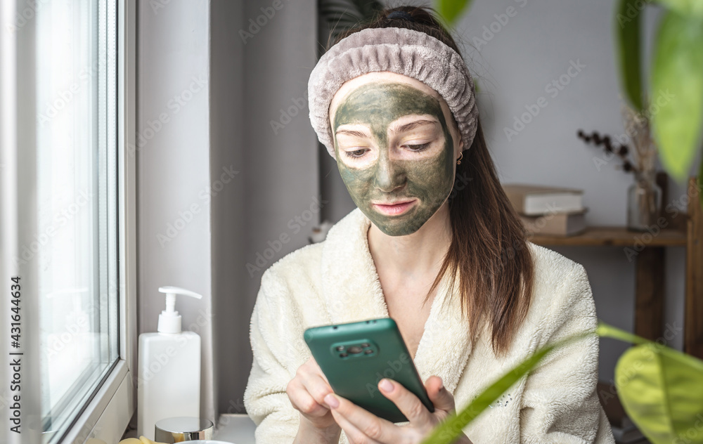 Young beautiful woman in a bathrobe and with a green cosmetic mask on her face is using her mobile phone Young beautiful woman in a bathrobe and with a green cosmetic mask on her face is using her mobile phone
