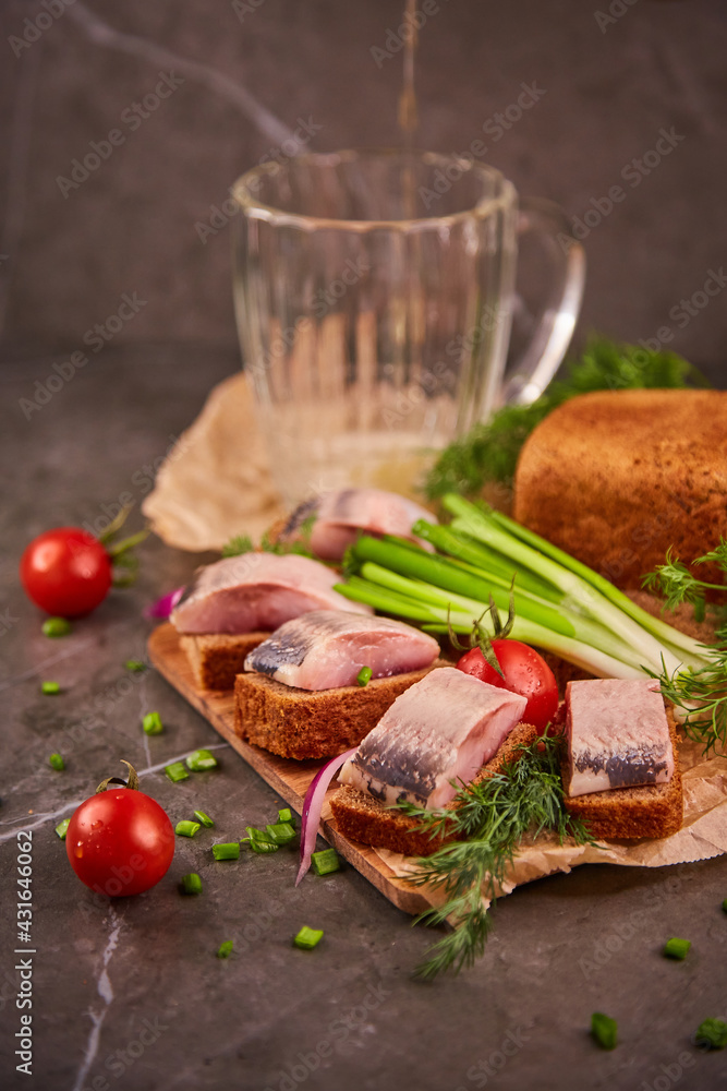 Slices of salted herring on black bread, light beer and fresh vegetables on a wooden kitchen board on paper. Delicious beer snack