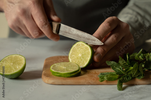 Bartender is cutting a juicy green lime on a chopping board with a knife, and a sprig of mint is lying next to it.