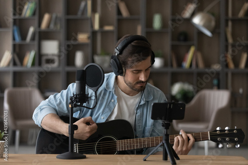 Man in headphones playing acoustic guitar, recording video on smartphone standing on tripod, using professional microphone, blogger or music teacher shooting course in home studio, sitting at desk
