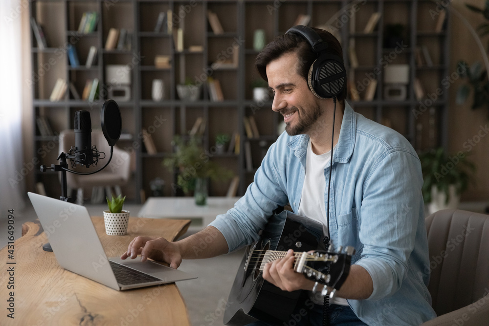 Smiling man in headphones using laptop, playing acoustic guitar, sitting at desk in home studio, happy artist musician making music, songwriting, student practicing, watching online course lesson Smiling man in headphones using laptop, playing acoustic guitar, sitting at desk in home studio, happy artist musician making music, songwriting, student practicing, watching online course lesson