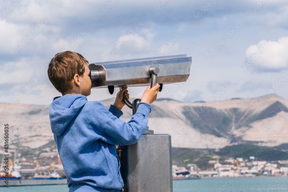 A blonde boy looks through a city telescope against a background of mountains and the sea