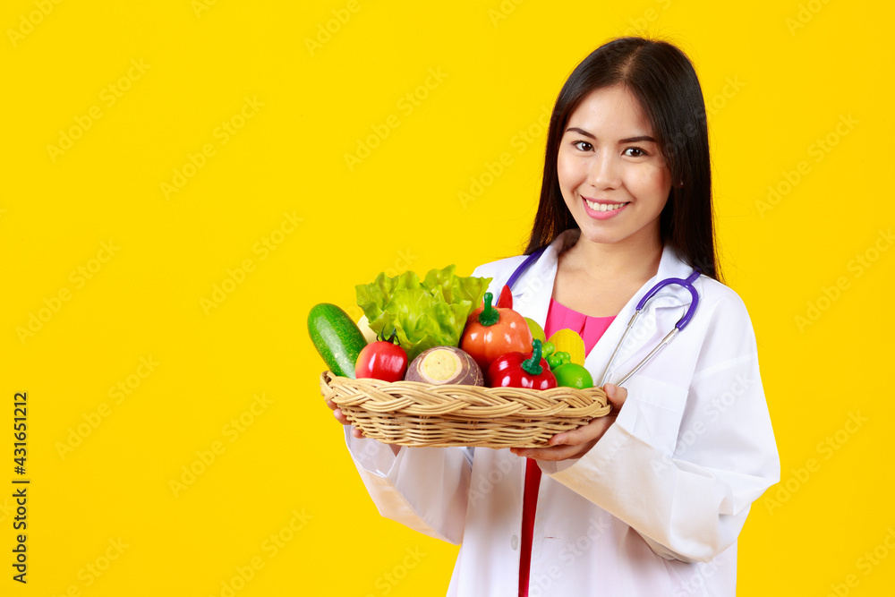 Asian young nice female doctor wearing white gown uniform with stethoscope, smiling and holding basket of vegetable and fruits, presenting nutrition, vitamin, healthy with isolated yellow background. Asian young nice female doctor wearing white gown uniform with stethoscope, smiling and holding basket of vegetable and fruits, presenting nutrition, vitamin, healthy with isolated yellow background.